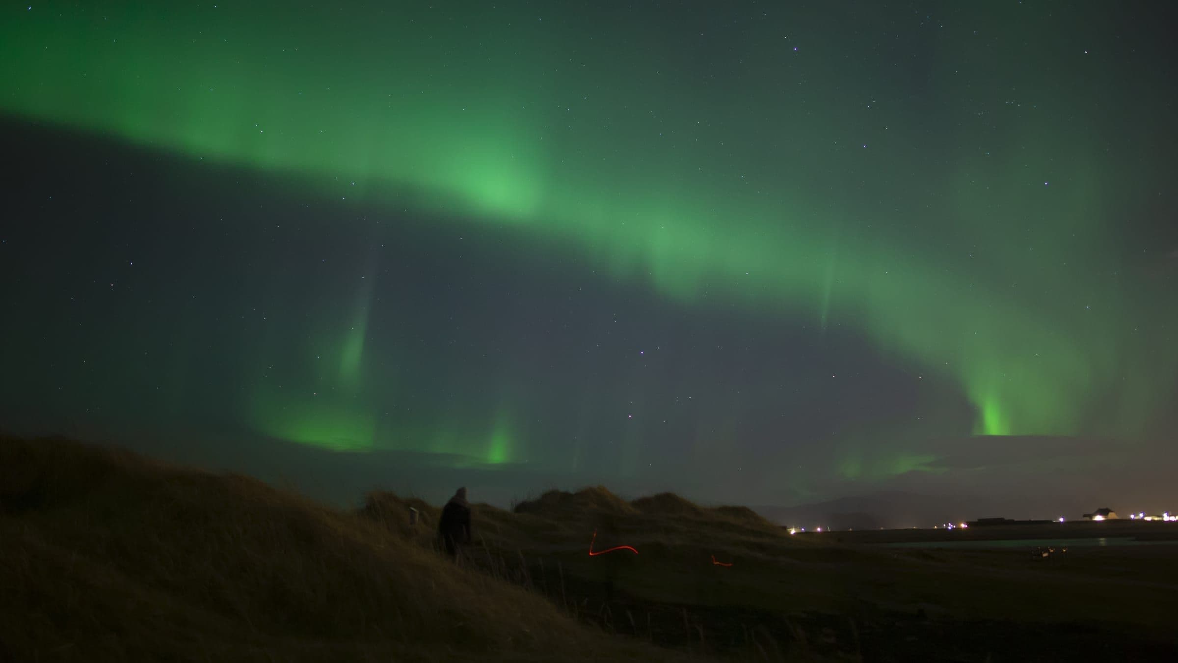 Aurora boreal verde extendida sobre dunas de pasto seco en Islandia, con una persona abrigada de pie mirando el cielo.