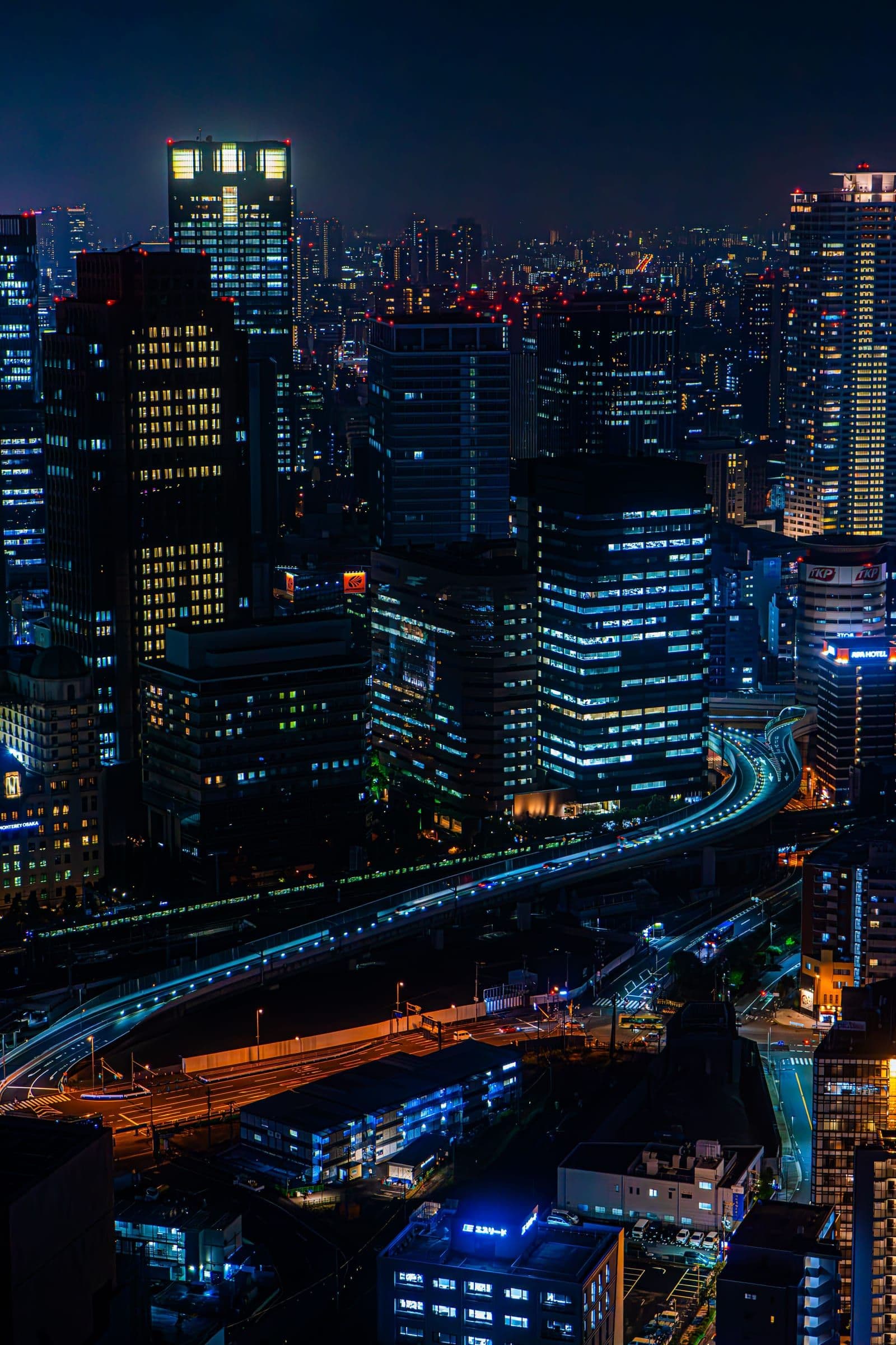 Skyline nocturno de Osaka visto desde lo alto, con rascacielos iluminados y las estelas de luz de una vía elevada.