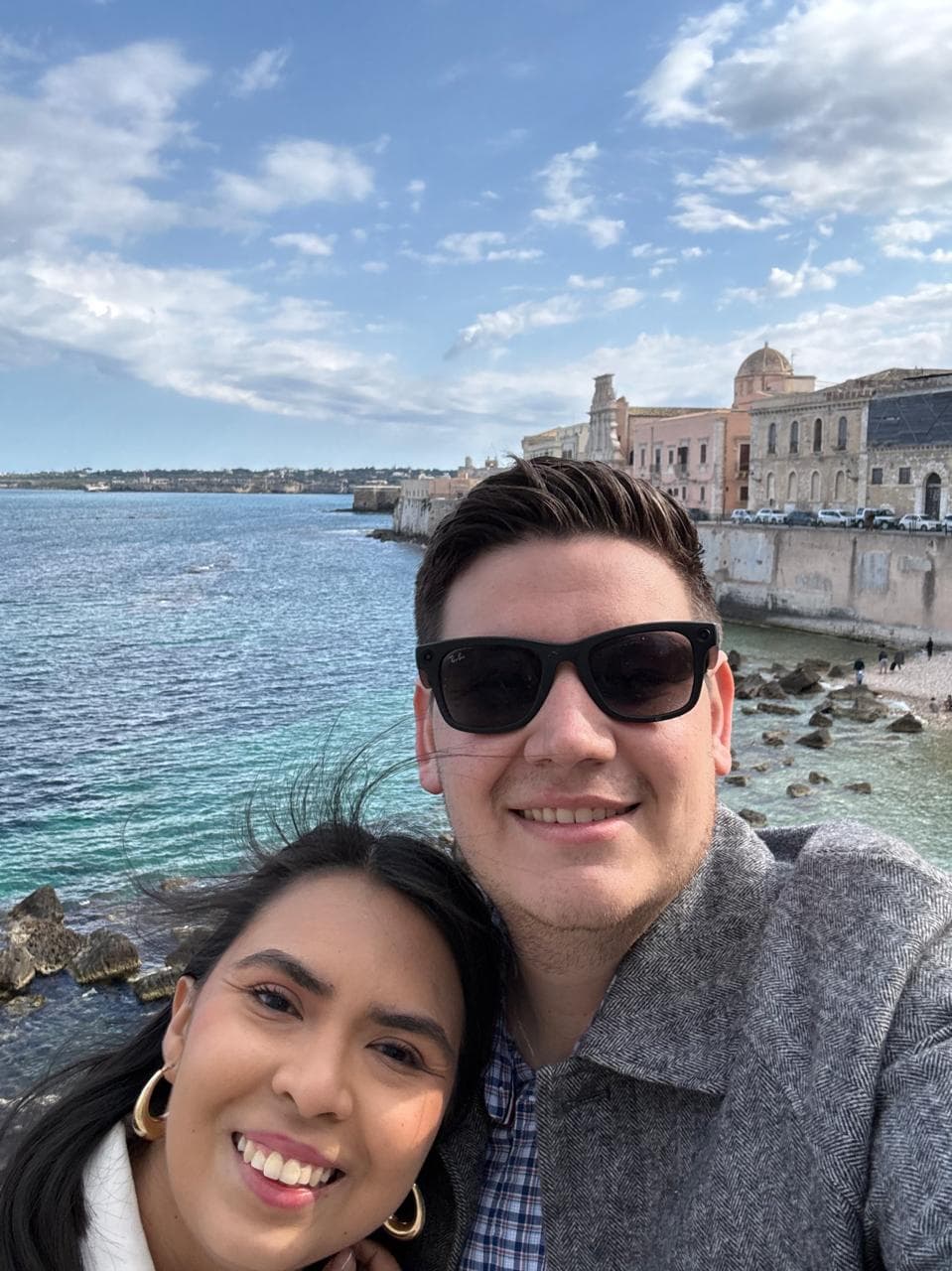 Sergio y Alexia sonriendo junto al mar en Ortigia, Siracusa, con los edificios color pastel del malecón detrás.